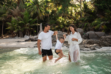 Wall Mural - Happy smile european family have rest and run on white sand beach. Father, long black chestnut hair mother, blond baby boy. white cotton clothes. boho dress. t-shorts.Thailand. Aquamarine crystal sea