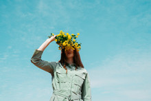 Redheaded Young Woman Against Sky Covering Eyes With Bunch Of Yellow Flowers