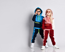 Little Kids, Boy And Girl, In Sunglasses And Hoods, Colorful Tracksuits, Sneakers. They Posing Isolated On White Studio Background