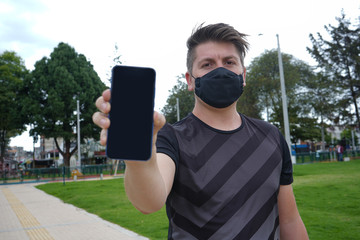 blond man in a park outdoors showing the screen of his cell phone with masks