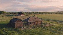 Abandoned Deserted Wooden Farmhouse With A Windmill And Outbuildings On The Background. Old Farm Buildings On A Green Field At Summer Sunset. Aerial View 