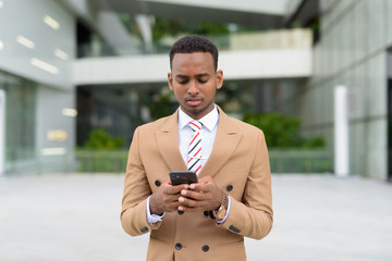 Wall Mural - Young handsome African businessman using phone in the city outdoors