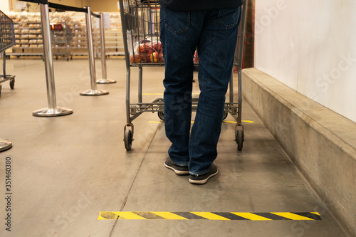 A man waiting in queue with cart while doing grocery shopping in a supermarket. Social Distance Shopping Line Up. Epidemic and coronavirus protection measures. Yellow and black tape on store\'s  floor.