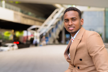 Wall Mural - Happy young handsome African businessman smiling and sitting in the city outdoors