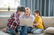 © zinkevych - Blonde female sitting between curly boy and dark-haired girl on sofa, holding laptop on her knees