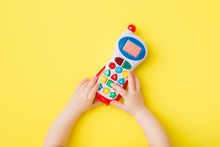 Baby hands playing with colorful toy of mobile phone on bright yellow table background. Closeup. Point of view shot. Top down view.