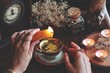 Wiccan witch wearing vintage jewelry holding yellow candle and pouring wax into a red gold vintage teacup as a divination. Reading Candle Wax - Carromancy, Ceroscopy among nature items dried flowers