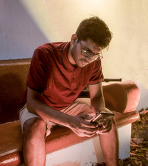 Stylish young guy in his early 20s, wearing casual attire of tshirt and shorts with eyeglasses, is using smartphone to access information from internet at free wifi zone, outdoors during night hours.