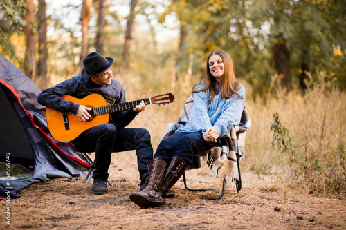 Fotografía Portrait of young couple sitting on camp chair with guitar near camp tent