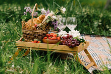 Picnic Basket With Strawberries, Grapes And Buns On The Green Grass In The Garden