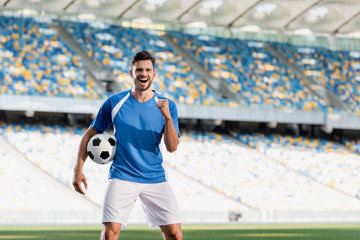 Wall Mural - happy professional soccer player in blue and white uniform with ball showing yes gesture on football pitch at stadium