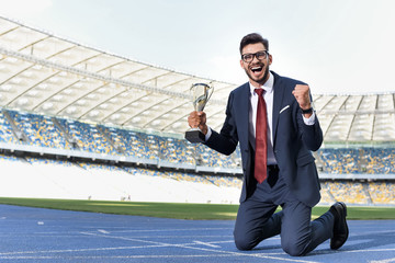 Wall Mural - happy young businessman in suit standing on knees on running track with trophy at stadium