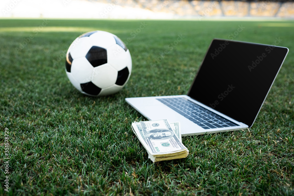 selective focus of soccer ball, money and laptop with blank screen on grassy football pitch at stadium, online betting concept - obrazy, fototapety, plakaty 