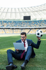 Wall Mural - young businessman in suit with laptop and soccer ball sitting on football pitch at stadium and yelling, sports betting concept