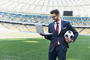 Wall Mural - smiling young businessman in suit with laptop and soccer ball at stadium, sports betting concept