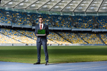 Wall Mural - smiling young businessman in suit showing laptop with online trade website at stadium