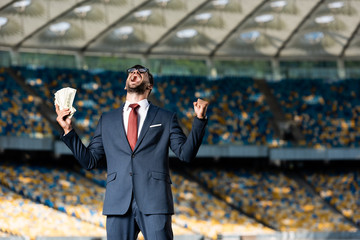 Wall Mural - young businessman in suit and glasses with money showing yeas gesture and yelling at stadium