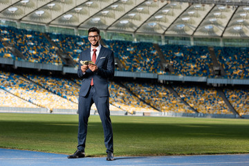 Wall Mural - smiling young businessman in suit and glasses with money at stadium