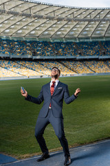 Wall Mural - young businessman in suit and glasses holding smartphone and showing yeas gesture at stadium, sports betting concept