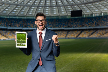 Wall Mural - cheerful young businessman in suit and glasses holding digital tablet with healthcare app and showing yes gesture at stadium