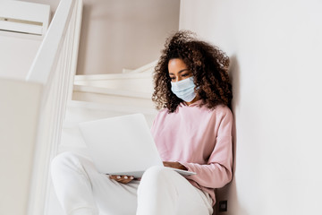 Poster - Curly African American freelancer in medical mask sitting on stairs and using laptop