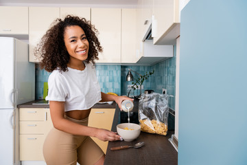 Wall Mural - happy african american woman pouring milk in bowl with cornflakes