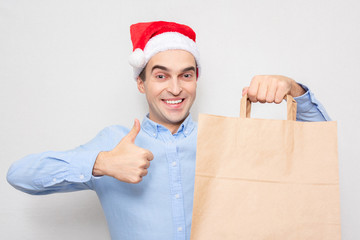 Wall Mural - Concept of a new year. Man in Santa Claus hat with gift bag, man shows thumb up, portrait, white background, copy space