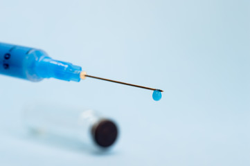 A close-up syringe needle with a drop of solution at the end against an empty vial on a blue background