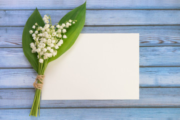 Wall Mural - bouquet of flowers lily of the valley and empty paper sheet on blue rustic table from above, white e