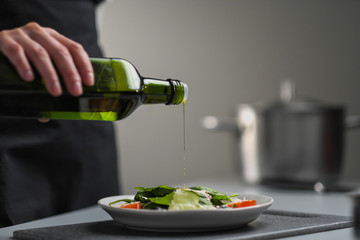 Wall Mural - A female chef in a white uniform and a black apron in the restaurant kitchen. Cooking. The cook pours olive oil from a green bottle.