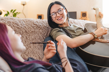 Smiling girl playing on guitar while her girlfriend smoking from pipe indoors