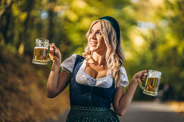 Wall Mural - Pretty happy blonde in dirndl, traditional festival dress, holding two mugs of beer outdoors in the forest