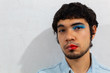 © Antonio Tanaka - Non-binary bearded young Hispanic man on a white background, with flamboyant makeup on one half of his face, red lips and blue eye shadows