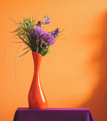 A bunch of purple violet spring flowers in an orange vase on warm yellow background. A colorful photo with free blank copy space for text. For cards, posters, website decoration