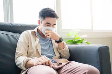 Fototapete - Asian man working from home wearing protective facemask sitting on sofa in living room feeling sick falling in illness coughing sad and tired, quarantine self isolation, coronavirus covid-19 pandemic