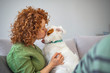 Close-up portrait of pleased girl with curly red hair embracing funny dog with eyes closed. Smiling young woman in green shirt enjoying good day and posing with pet on sofa at home.