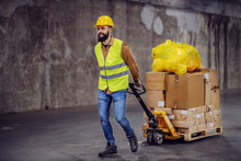 Full Length Of Smiling Hardworking Bearded Worker In Vest, With Safety Helmet On Head Pulling A Pallet Truck With Boxes, Sack And Building Material. Building In Construction Process Interior.