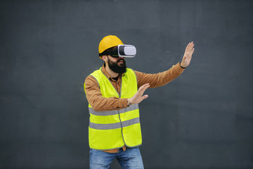 Wall Mural - Young bearded worker in vest, with safety helmet on head having vr goggles and enjoying virtual presentation.