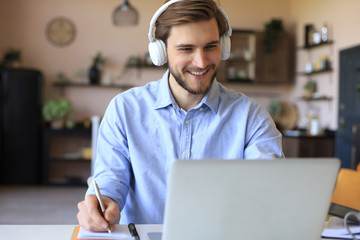 Confident businessman in earphones is writing notes or financial report while sitting at desk with laptop at home.