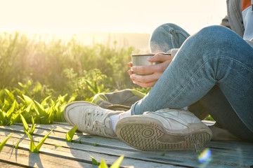 Close up of a young girl with a backpack is resting in a field on a wooden deck and drink hot tea from a steel thermos. Journey and outdoor concept.