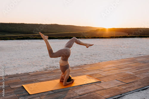 Young slim fit woman on a sport mat in yoga head stand pose with legs apart, at the sunset, outdoors. Sportive girl doing pilates.