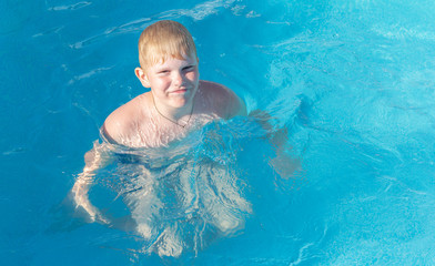 a strong blond boy with a smile swims in an outdoor pool. Child having fun in swimming pool. Summer vacation and healthy lifestyle concept