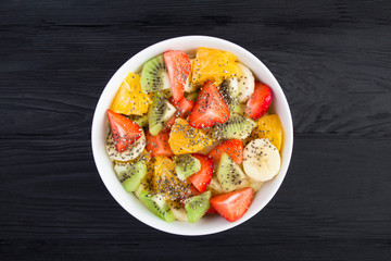 Wall Mural - Fruit salad with chia seeds in the white  bowl in the center of  the black wooden background. Top view. Closeup.