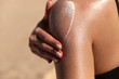 Leinwandbild Motiv Young woman applying sun cream or sunscreen on her tanned shoulder to protect her skin from the sun. Shot on a sunny day with blurry sand in the background