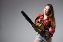 Sexy Girl In A Red Shirt, Denim Shorts With A Chainsaw In Her Hands. Woman Builder On A Gray Background