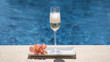 Glass with champagne with foam on swimming pool or sea blurred background. Sparkling white wine on tray with flower. Cold champagne, luxury alcohol drink, romantic vacation.