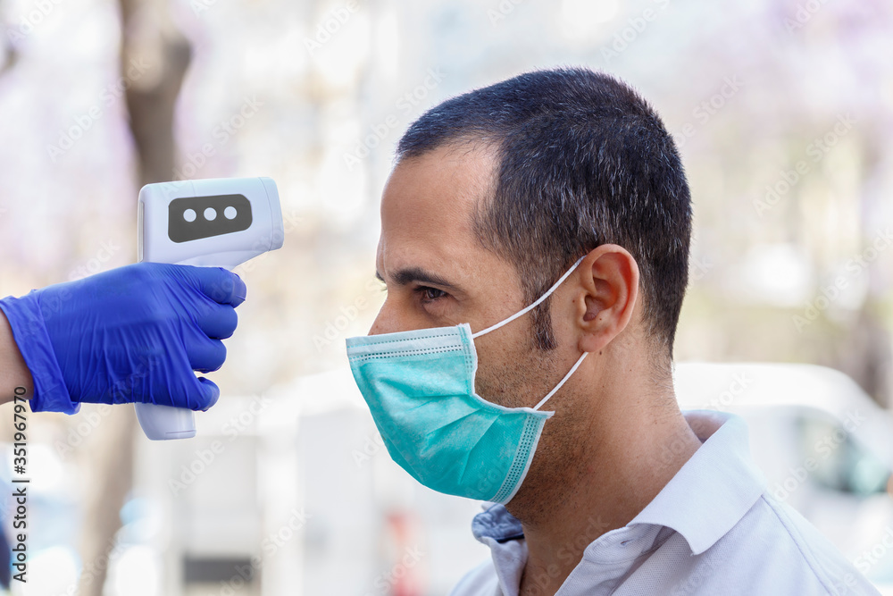 A uomo moro con mascherina chirurgica viene presa la temperatura del corpo con termometro elettronico digitale, isolato su sfondo urbano - obrazy, fototapety, plakaty 
