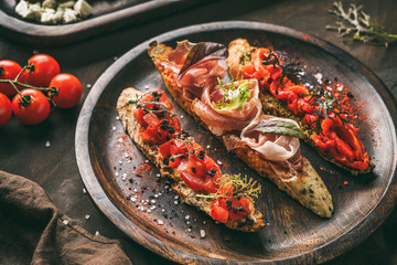 Canvas Print - Variety of bruschetta with prosciutto, tomatoes, parmesan cheese, sauce pesto, grilled pepper on ciabatta bread on wooden plate over dark background. Sandwiches set of wine, top view