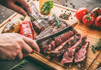 Wall Mural - Hands cut grilled tomahawk meat medium rare or rib eye steak on wooden cutting board with grilled vegetables on dark background, close up, toning