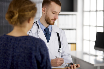 Friendly red-bearded doctor and patient woman discussing current health examination while sitting in sunny clinic. Medicine concept